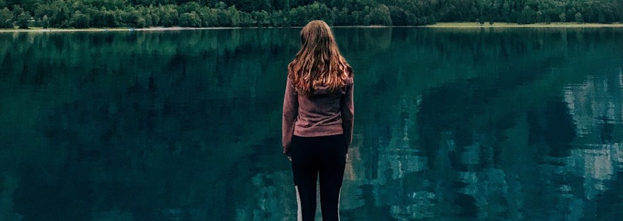 Young woman standing on a dock on a lake looking out at trees on a cliff face.